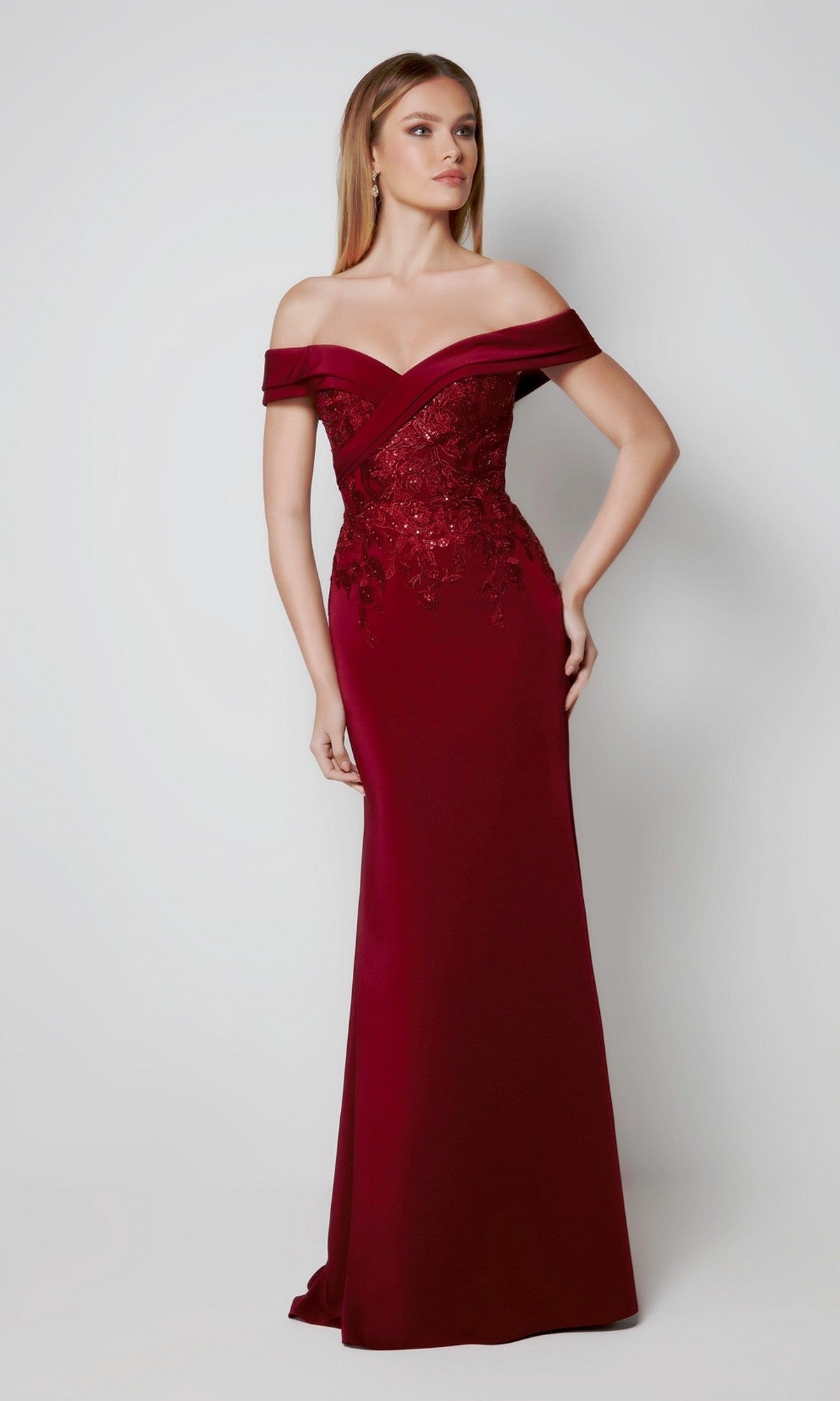 A woman wears the Alyce 27571 Long Formal Dress in burgundy, a floor-length off-the-shoulder gown with lace bodice detailing, made from stretch-crepe, standing against a plain light backdrop.